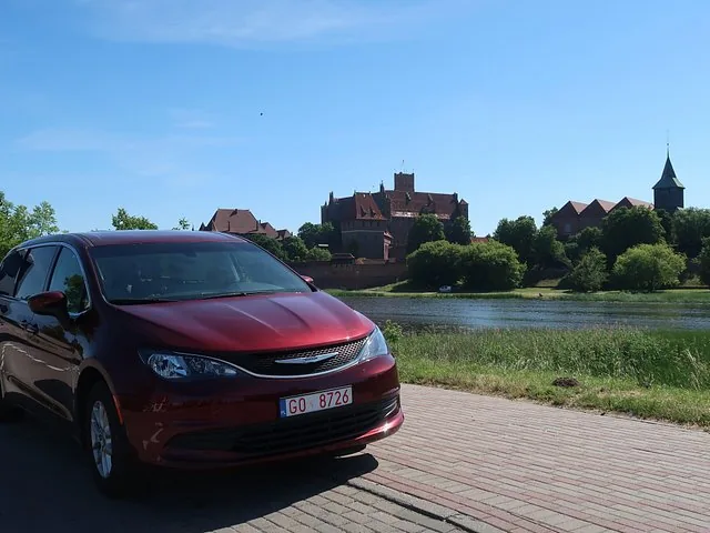Red minivan parked along the river with Malbork Castle in the distance.